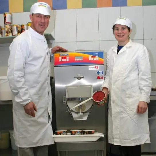A pair of farmers in cooking clothes smiling next to an ice cream machine.